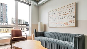 Een moderne woonkamer met een grijze bank, een houten salontafel en een bruine fauteuil. Een groot raam biedt uitzicht op stadsgebouwen.