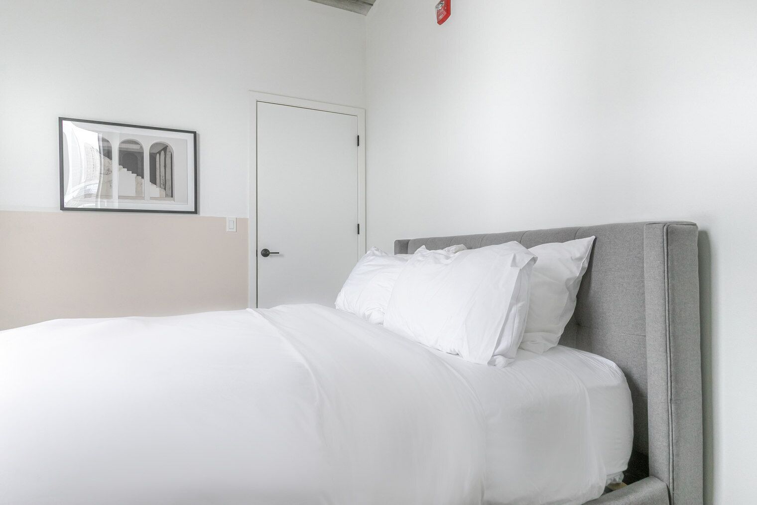 A neatly made bed with white bedding in a room with a grey headboard, a white door, and a framed picture on the wall.