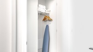 Ein schmaler Flur mit einer weißen Tür rechts, einem blauen Bügelbrett in der Mitte und einem Regal mit gefalteten weißen Handtüchern darüber.