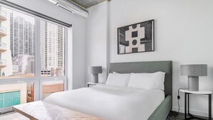 Ein modernes Hotelzimmer mit einem großen Bett, zwei Nachttischen mit Lampen, Blick auf hohe Gebäude durch große Fenster und einem geometrischen Kunstwerk an der Wand.