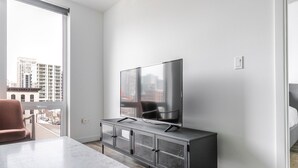 Een moderne woonkamer met een grote flatscreen televisie op een kast, een eettafel en een stadsgezicht door een groot raam.