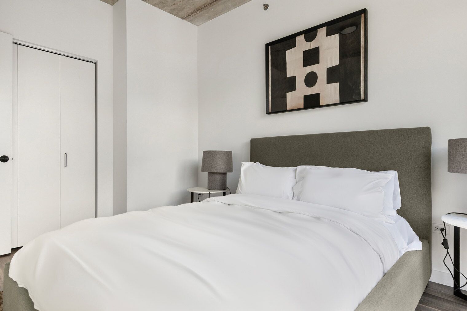 A modern bedroom with a large bed, a grey headboard, a bedside table with a lamp, and a wall-mounted abstract art piece.