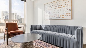 Un salon moderne avec un canapé gris, un fauteuil marron et une table basse ronde. Un grand tableau encadré est accroché au mur, et on peut apercevoir un paysage urbain à travers la fenêtre.