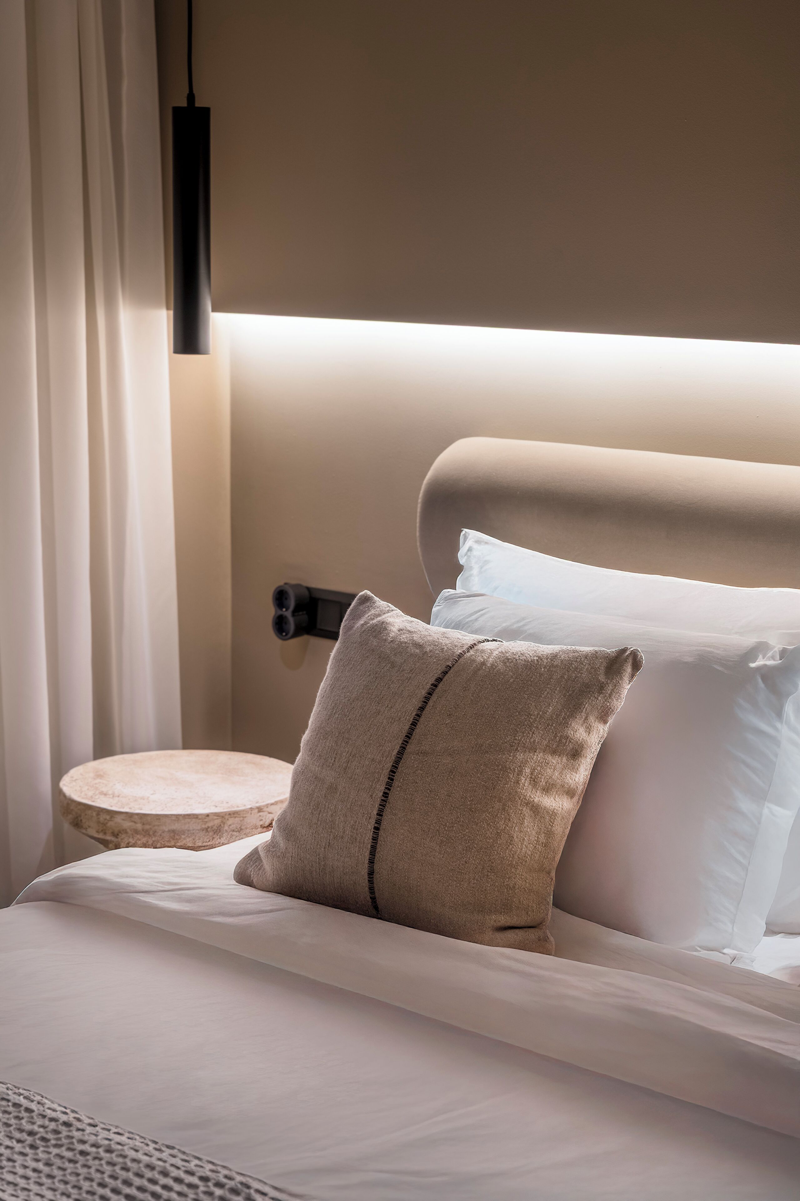 Een opgemaakt bed met witte lakens en een beige kussen, een rond nachtkastje en een zwarte hanglamp.