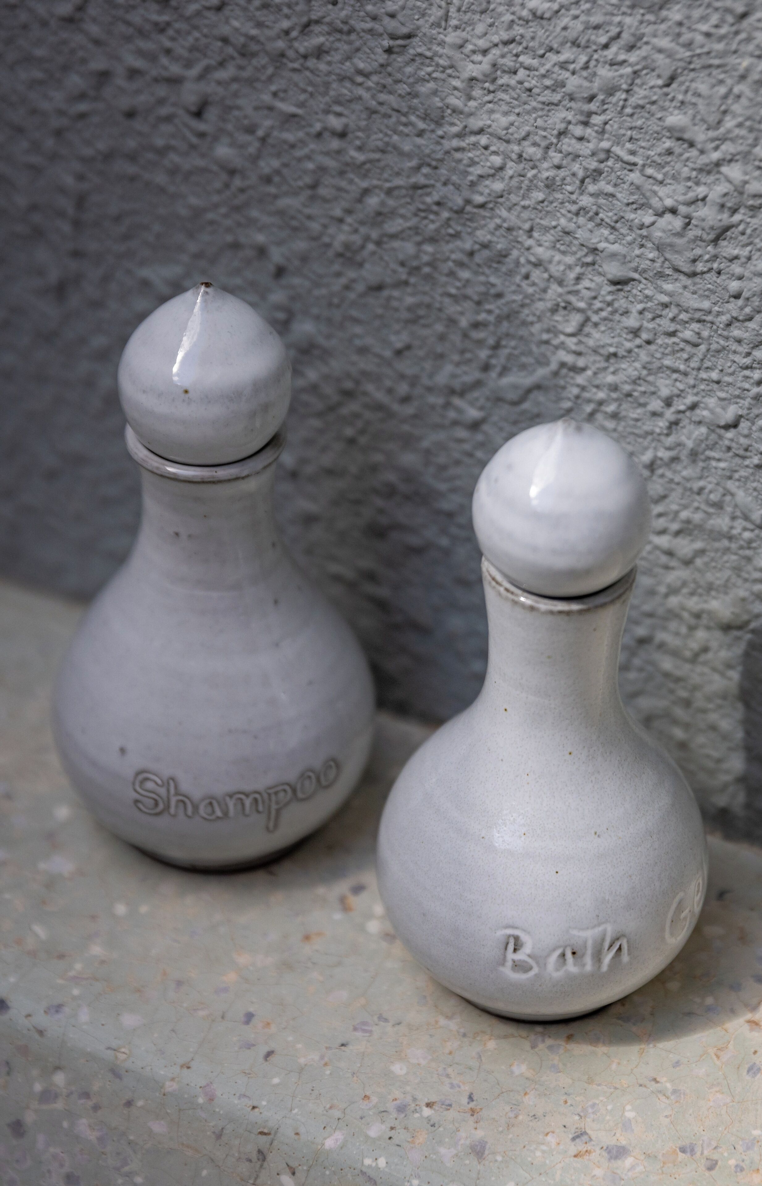 Two ceramic bottles labeled 'Shampoo' and 'Bath Gel' on a textured surface.