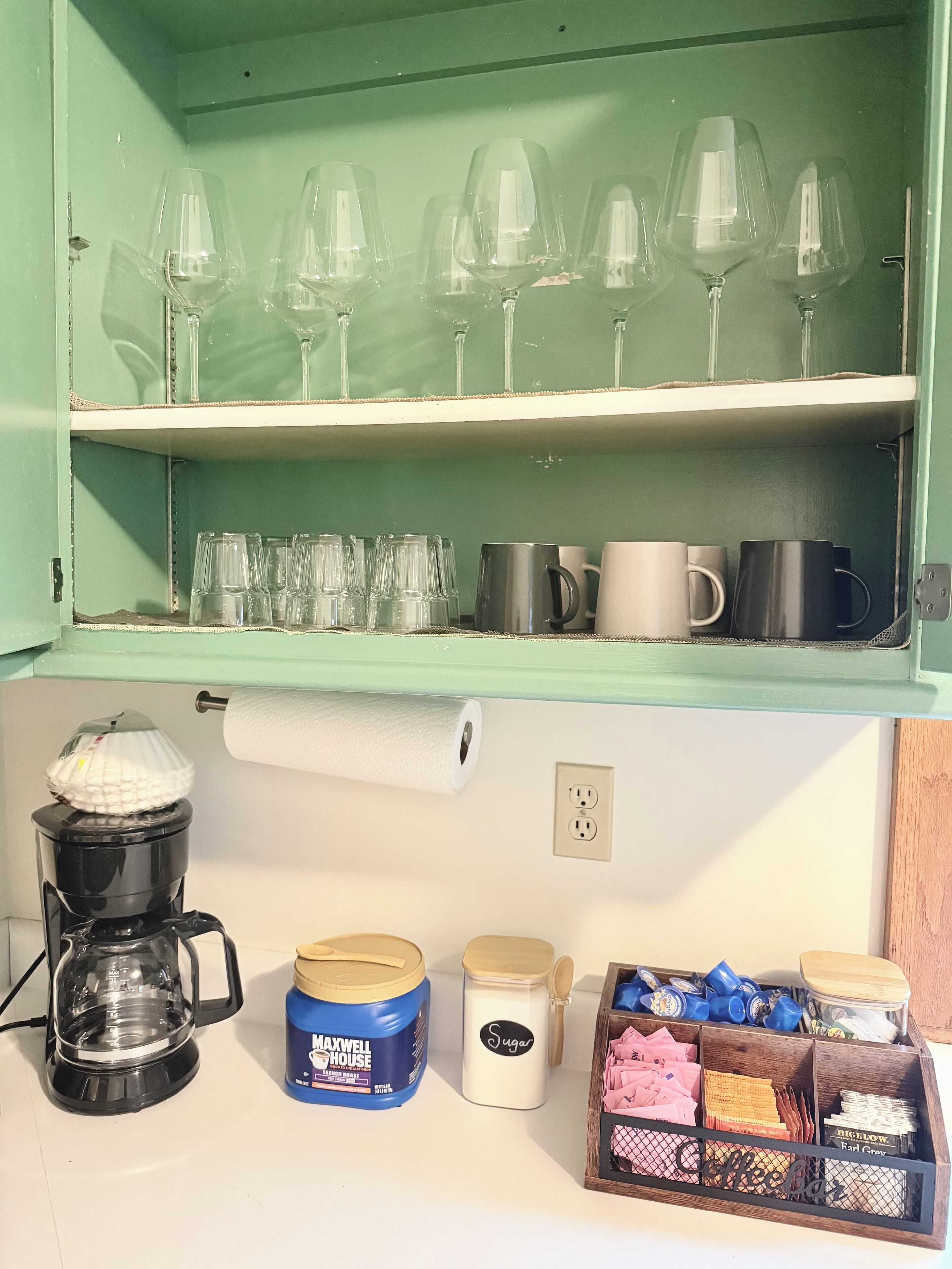 Una cucina con un armadietto verde che contiene bicchieri da vino, tazze e una macchina del caffè. Sotto l'armadietto, ci sono barattoli etichettati 'Maxwell House French Roast' e 'Zucchero'. Si vede anche una scatola di legno con diversi oggetti da cucina.