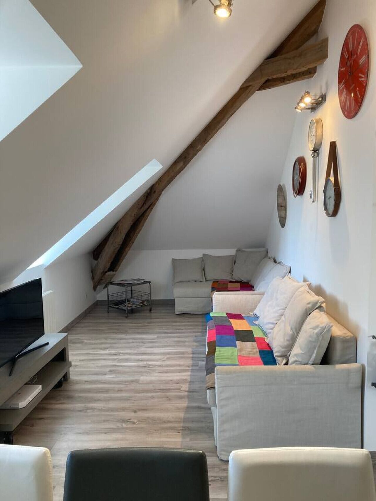 Een loft-achtige woonkamer met een schuin dak, zichtbare houten balken, een flatscreen televisie, een bank met een kleurrijk quilt en verschillende wandklokken.