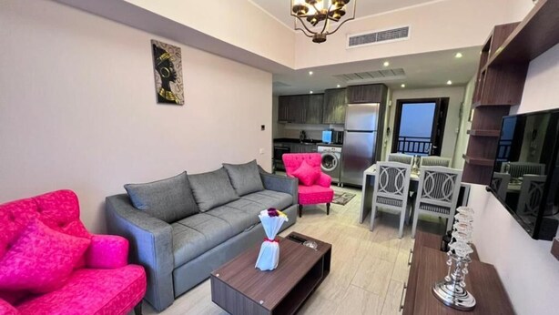 A modern living room with a grey sofa, a pink armchair, and a coffee table. There is a kitchen in the background with dark cabinets and a refrigerator. - Al Dau Heights Apartments (Hurghada)