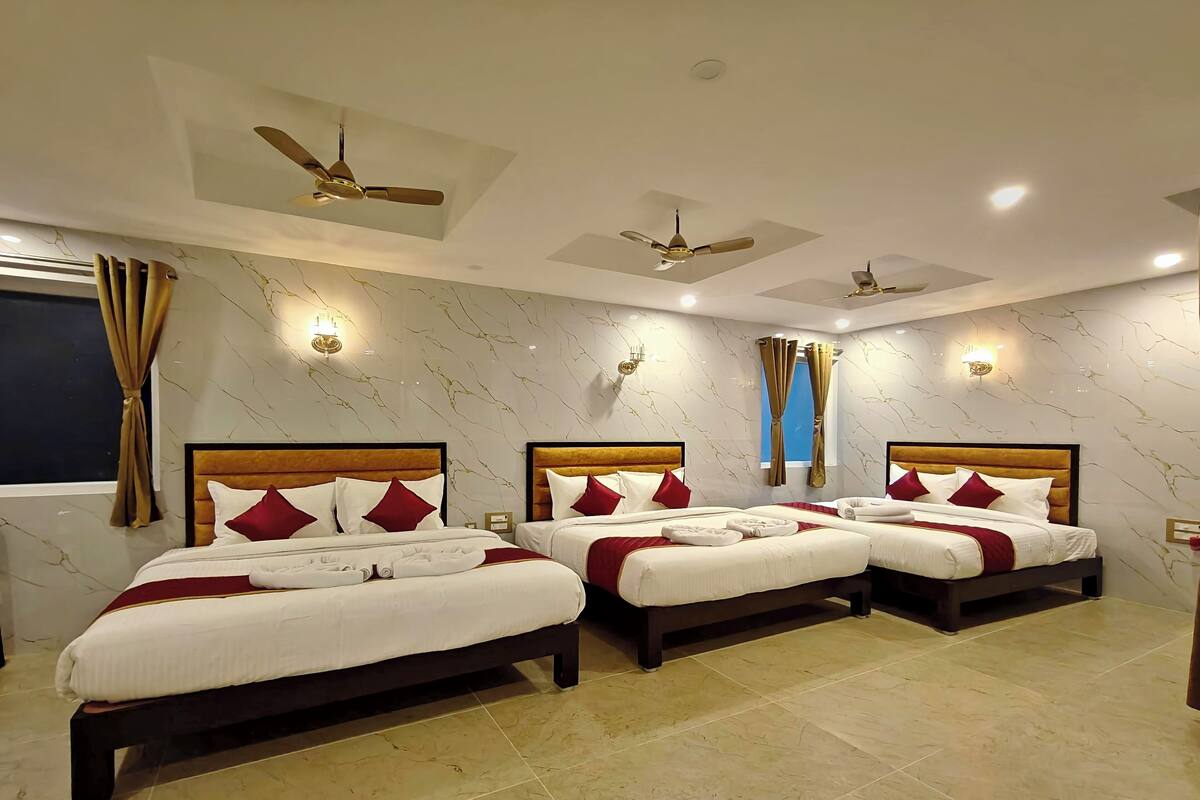 Habitación de hotel con tres camas, cada una con cabecero de madera y ropa de cama blanca y roja. Hay ventiladores de techo, apliques de pared y un ventanal con cortinas.