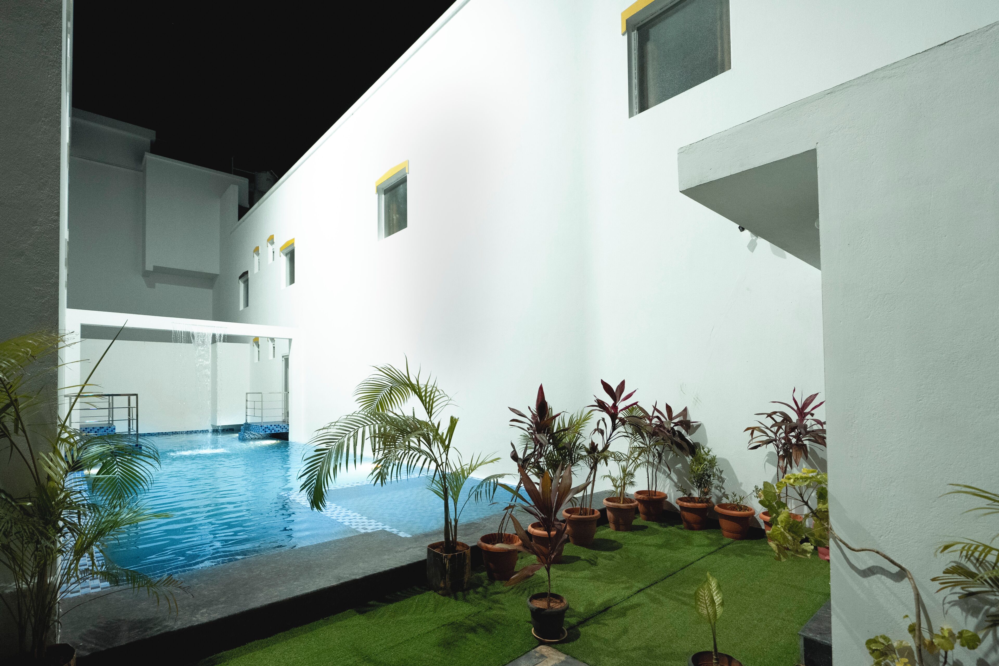 A swimming pool with a small waterfall feature, surrounded by potted plants and a green artificial turf area.