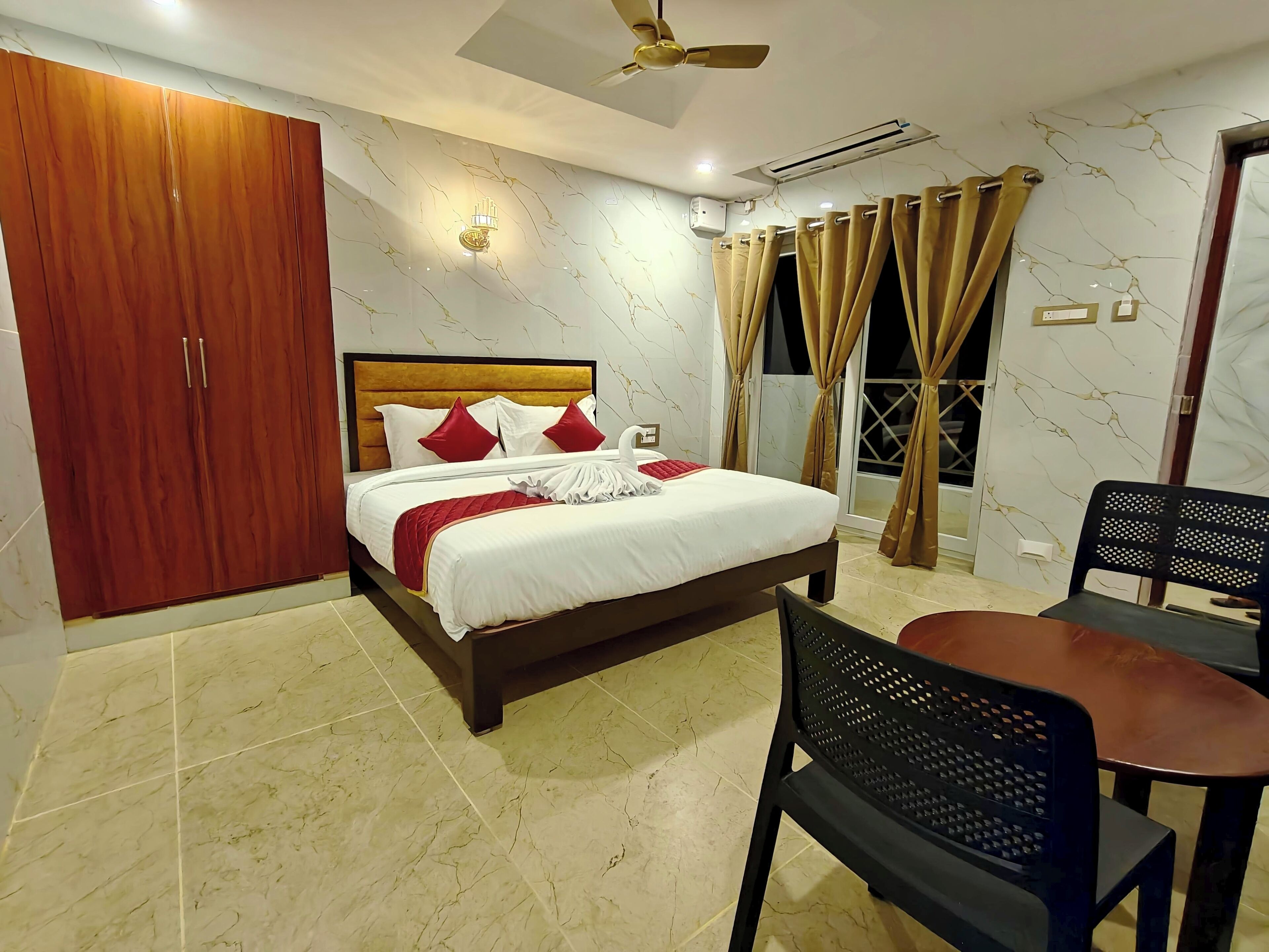 A bedroom with a bed, a wardrobe, a ceiling fan, an air conditioning unit, a dining table with chairs, and a view through the window.