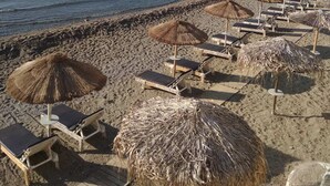 Ein Strand mit Liegestühlen und Reetsonnen schirm, freier Blick auf das Meer, und Gebäude an einem Hügel im Hintergrund.
