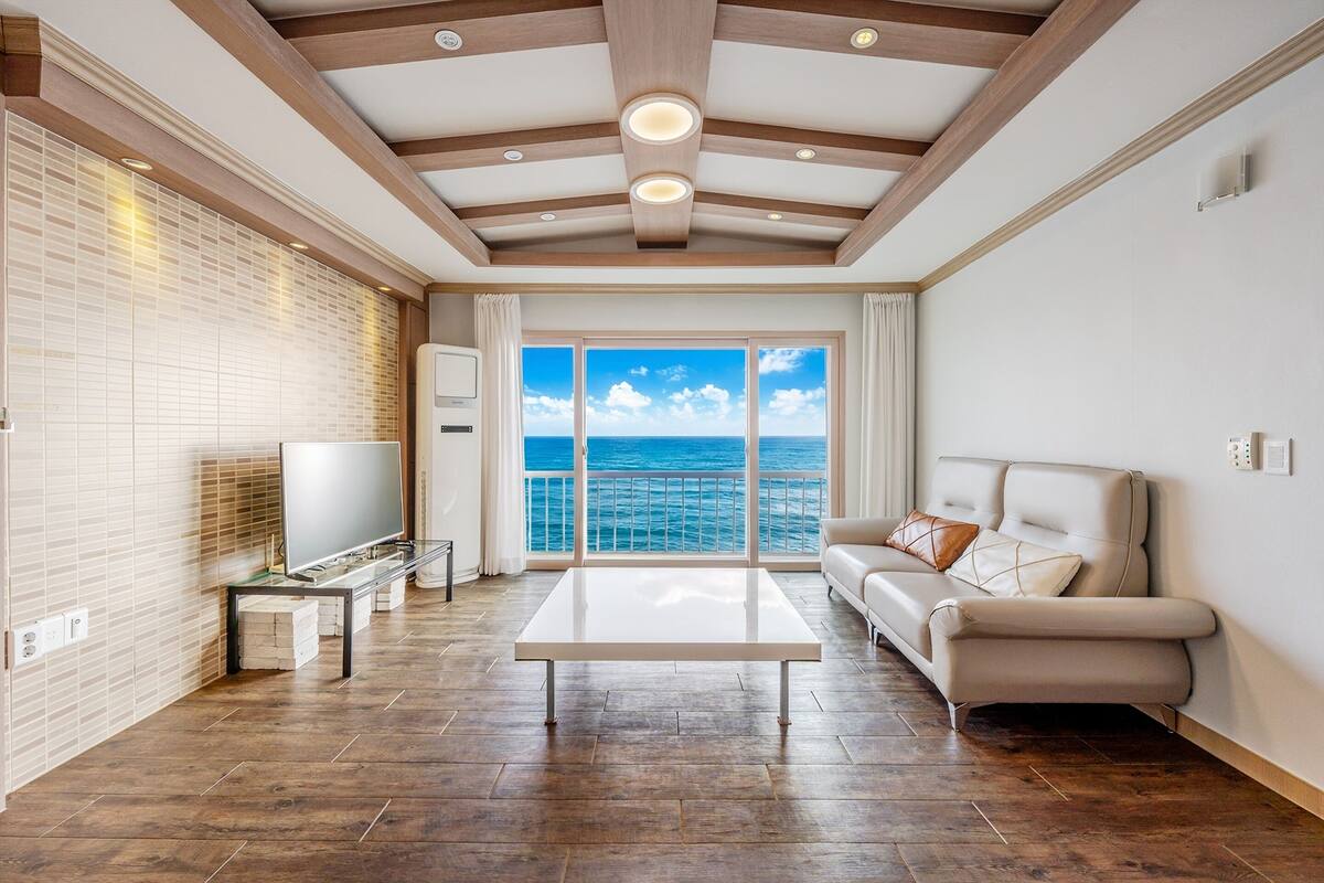 Un salon spacieux doté d’une grande fenêtre offrant une vue sur la mer, d’un téléviseur à écran plat, d’un canapé et d’une table basse.