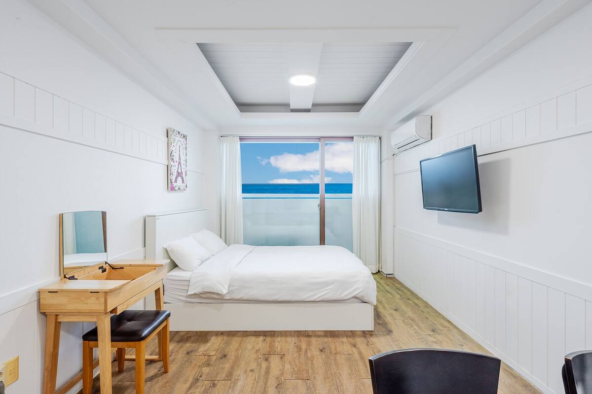 Une chambre à coucher comprenant un lit, un bureau, une chaise, un téléviseur et une fenêtre donnant sur la mer.