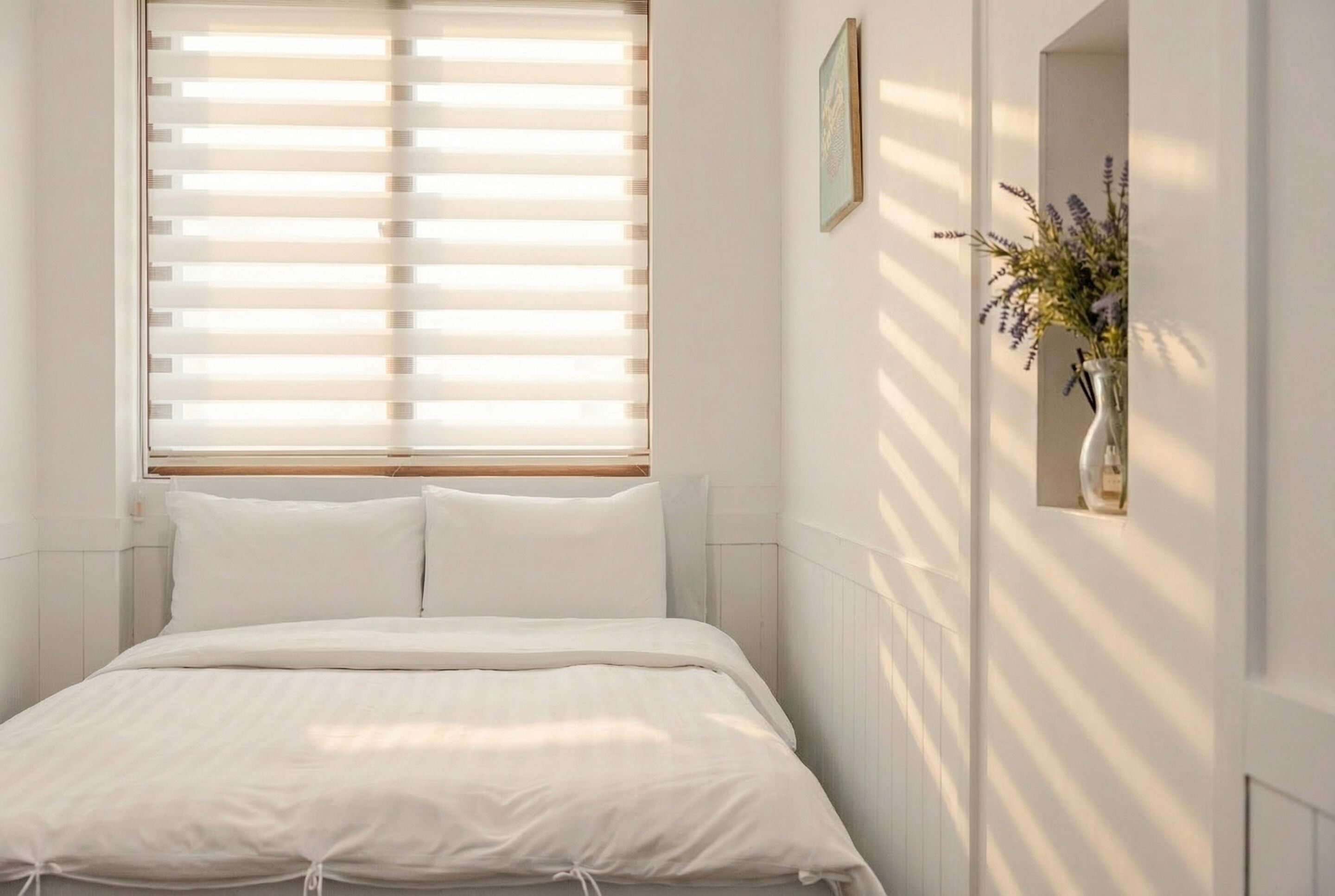 Un dormitorio con cama, paredes blancas, una ventana con persianas y un florero con flores en la pared.