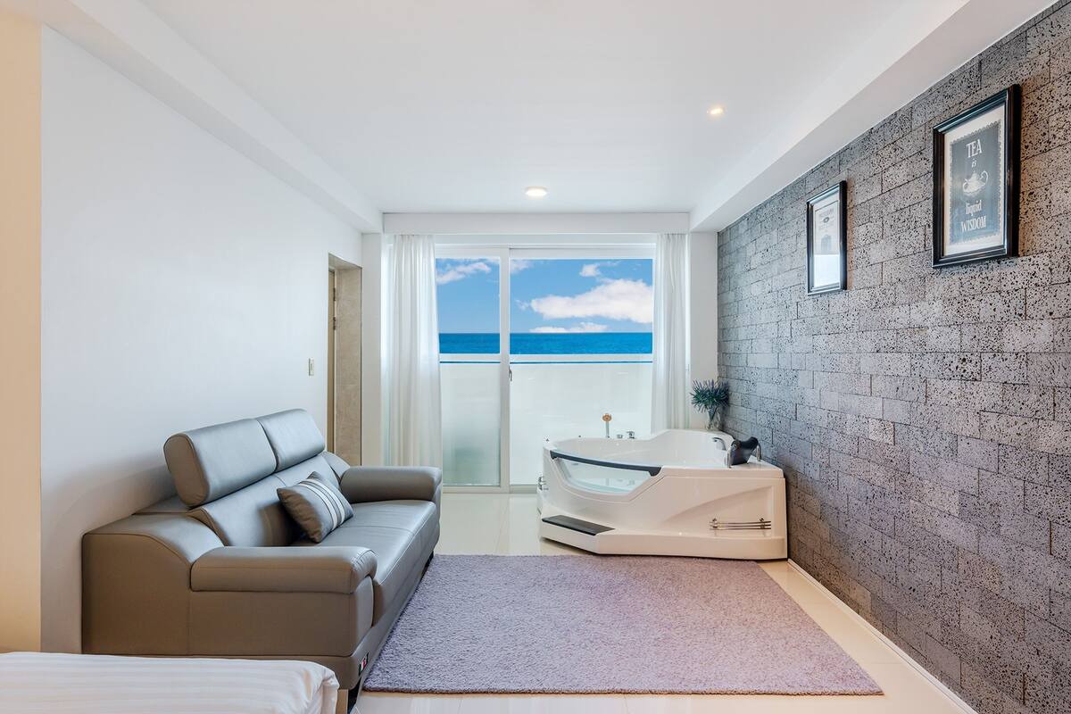 Une chambre moderne avec un mur en briques grises, un canapé gris, une baignoire blanche et une vue sur l’océan.