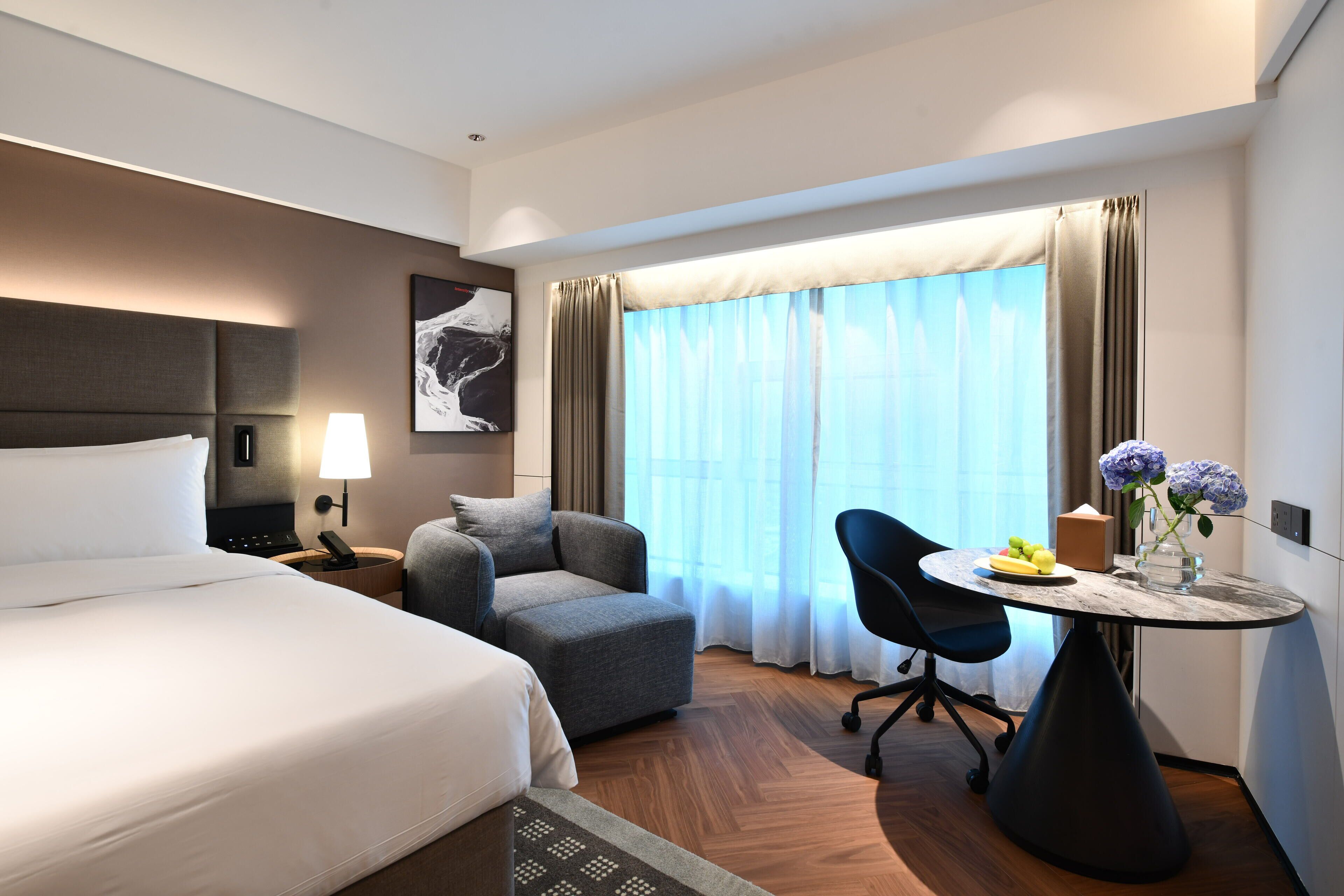 A hotel room with a bed, a desk, a chair, a table with flowers, and a window with curtains.
