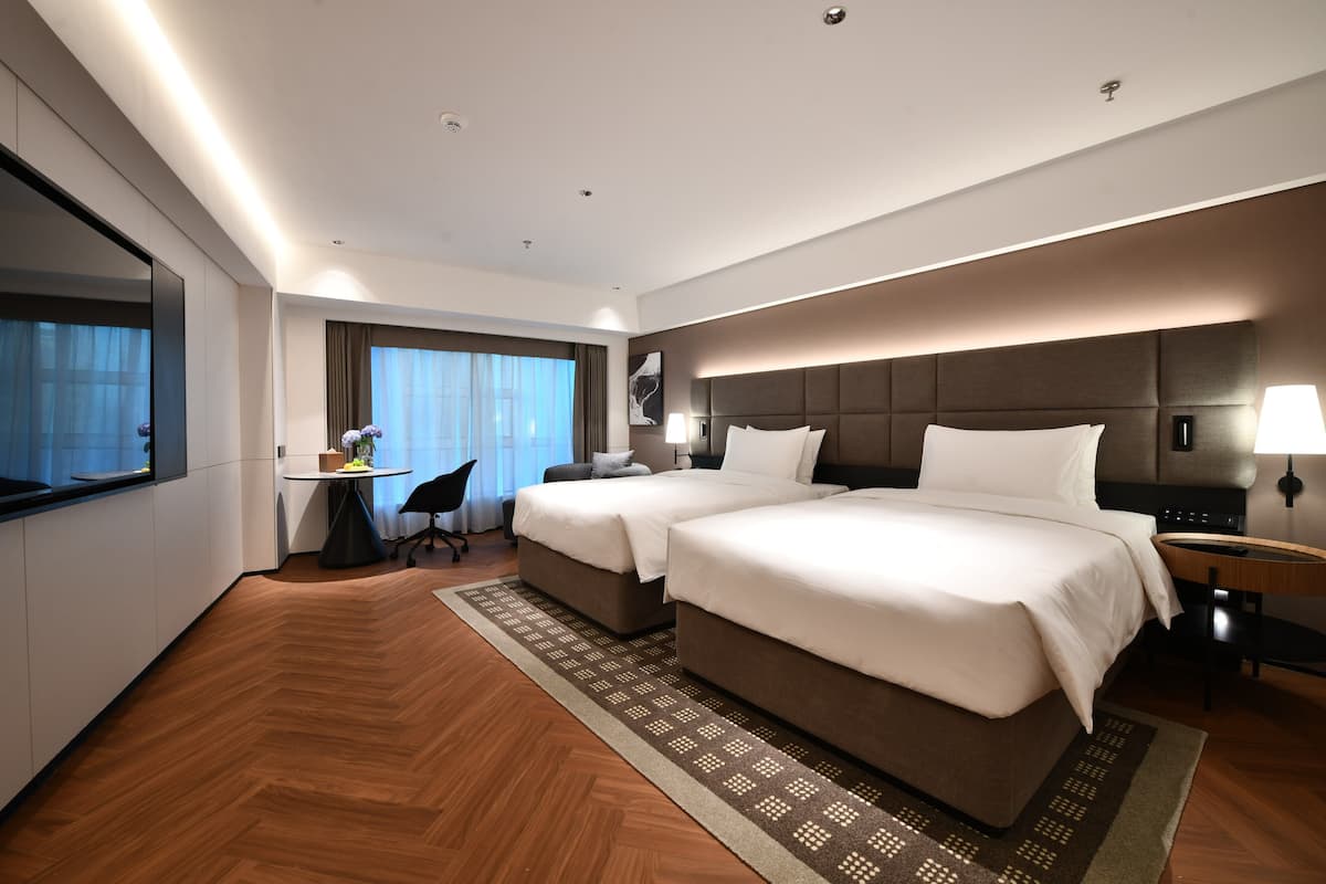 A modern hotel room with two beds, a desk, a chair, and a large window with curtains.