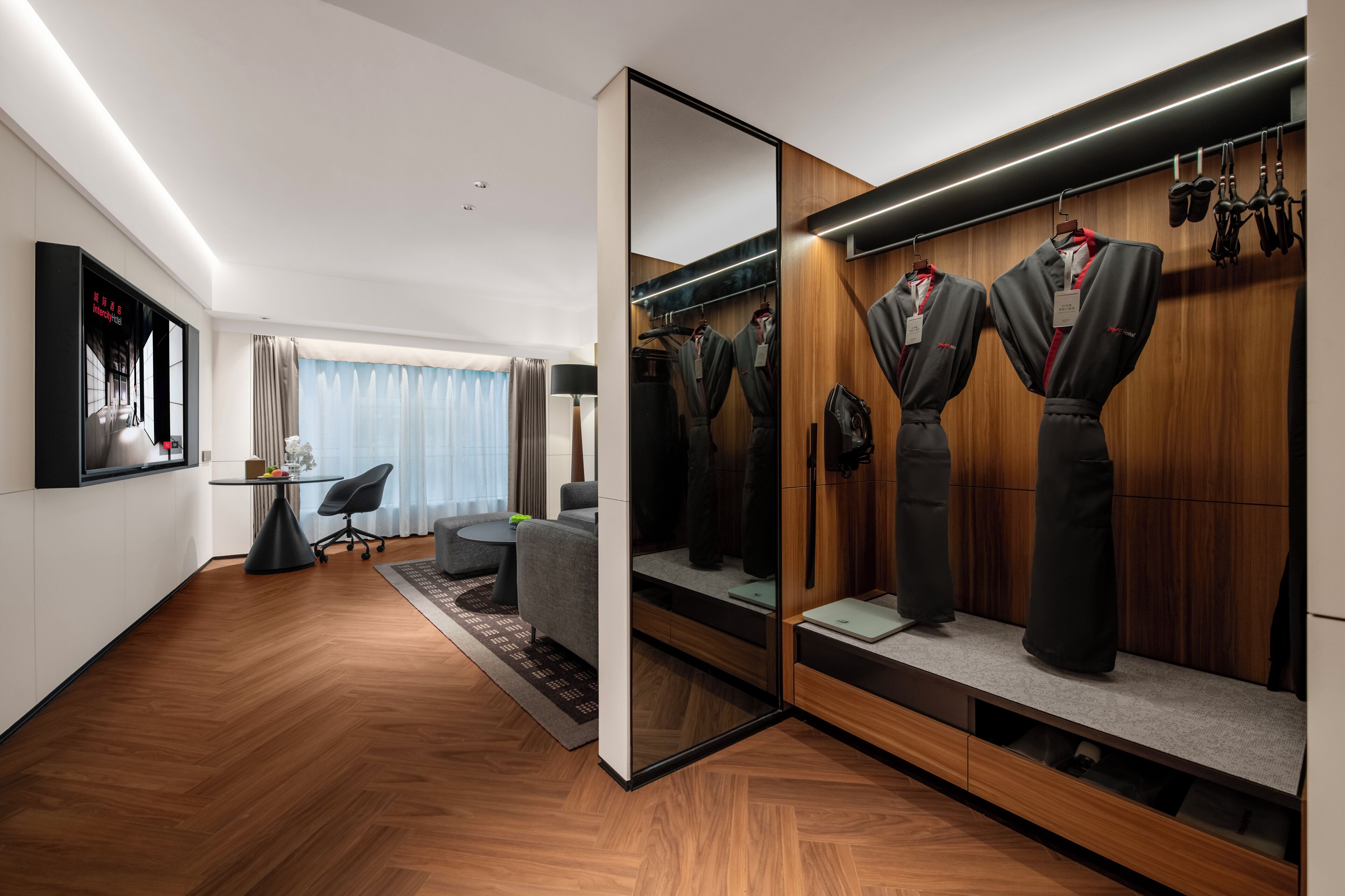 Ein modernes Hotelzimmer mit Holzboden, einer Sitzecke mit Sofa und Sesseln, einem Schreibtisch mit Lampe und einem begehbaren Kleiderschrank mit Kleidung und Schuhen.