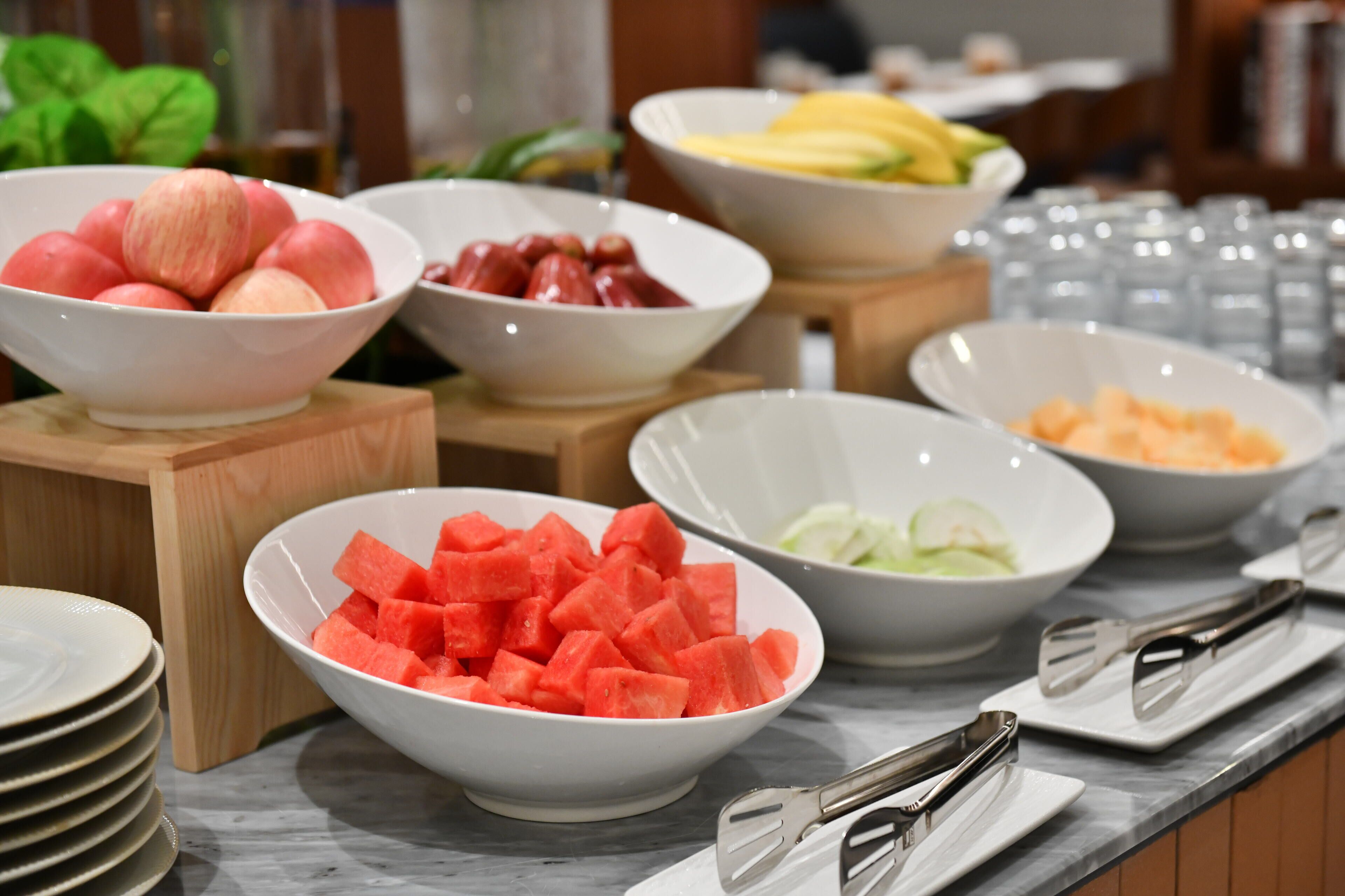 A buffet table with various bowls of fruits and vegetables, including apples, grapes, watermelon, and cucumbers.
