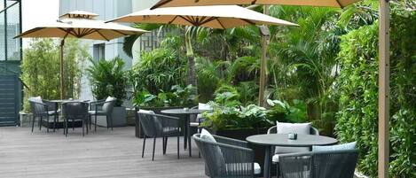 An outdoor patio area with round tables and chairs, surrounded by lush greenery and large umbrellas.
