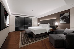 A modern hotel room with a large bed, a sofa, a desk with a lamp, a TV, and a sliding door closet.