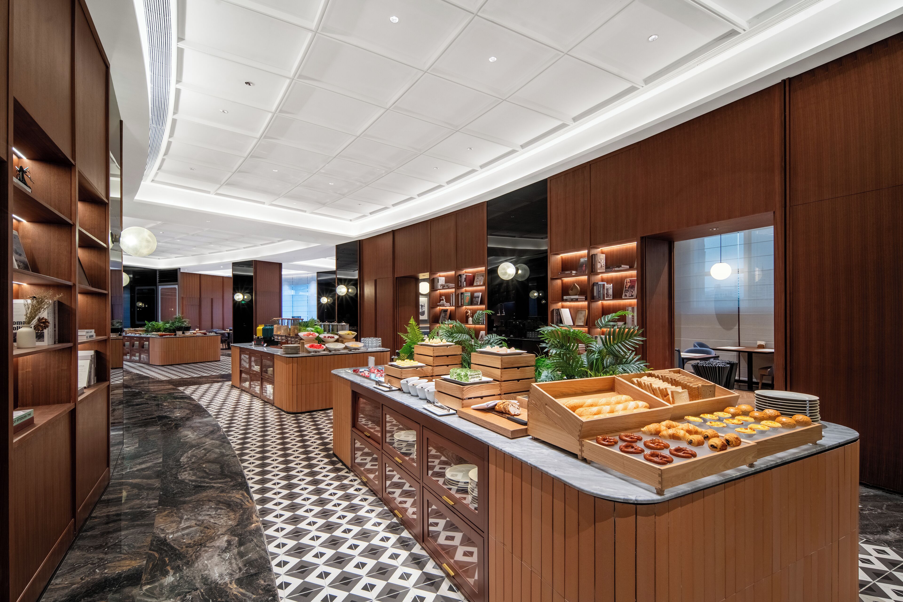 Un moderno vestíbulo de hotel con piso de mármol, paneles de madera y una zona de bufé con diversas opciones de comida.