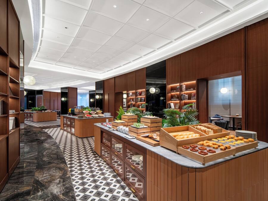 Un moderno vestíbulo de hotel con piso de mármol, paneles de madera y una zona de bufé con diversas opciones de comida.