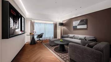 A modern hotel room with a grey sofa, a round coffee table, a TV mounted on the wall, and a large window with curtains.