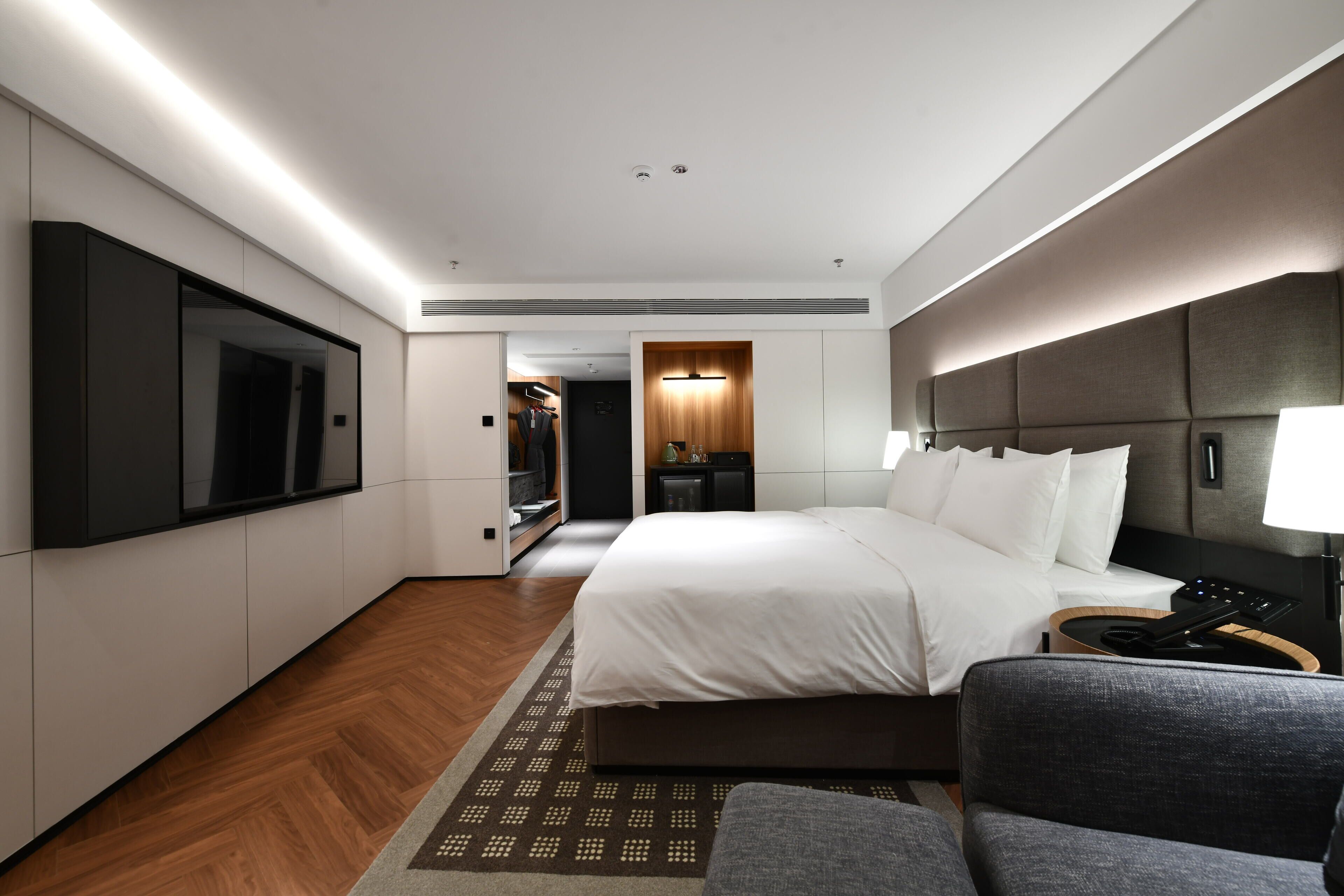 Una habitación de hotel moderna con una cama grande, un televisor de pantalla plana en la pared, un espacio de descanso con una silla y una mesita con una lámpara.