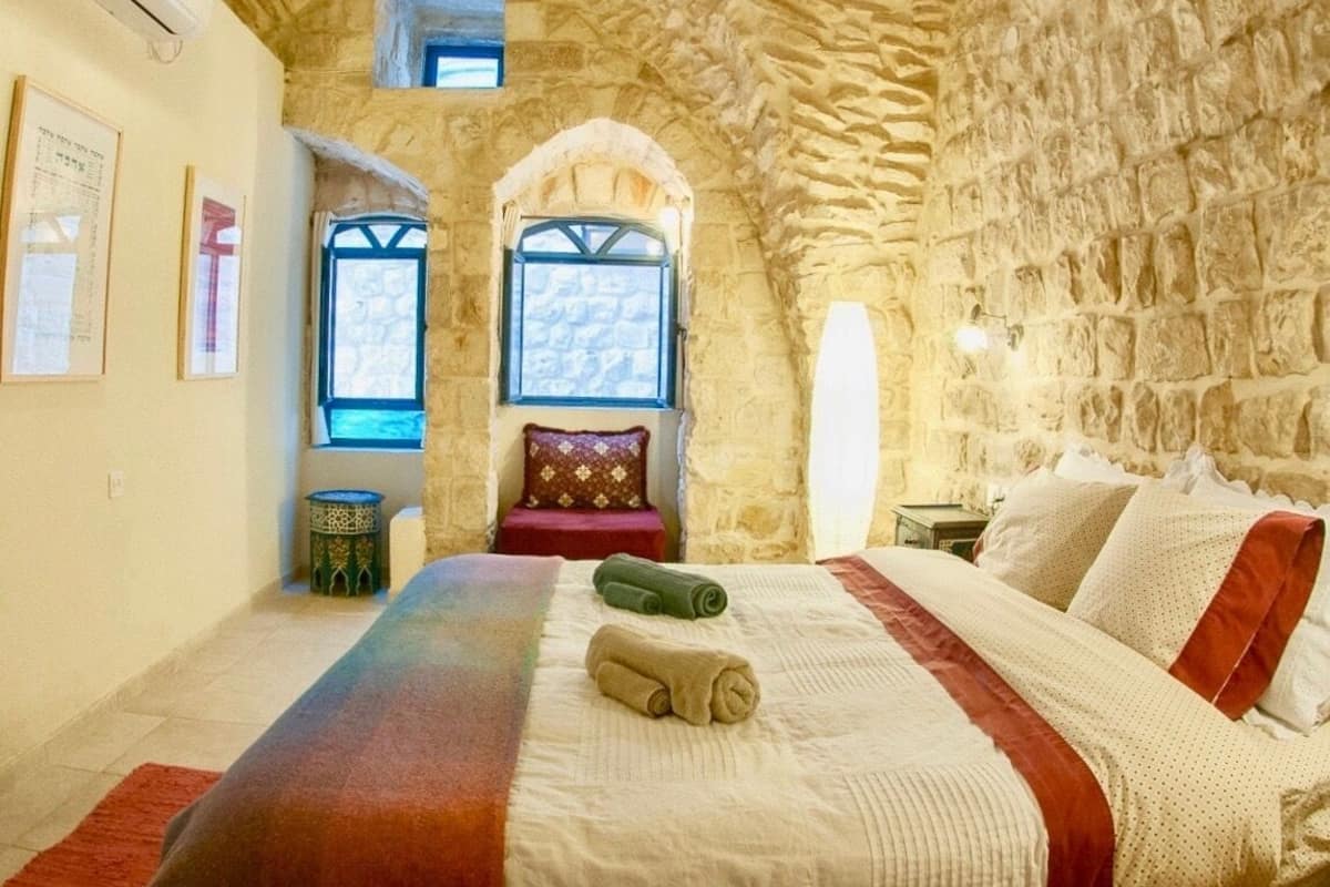 A bedroom with stone walls, a bed with a red and white blanket, a small table with a green lamp, and two windows with arched tops.