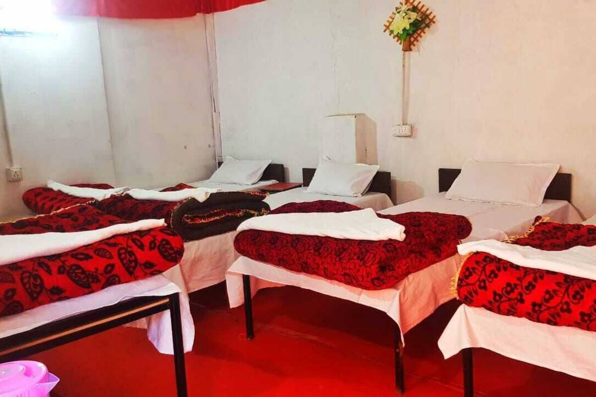 Une chambre d’hôtel avec quatre lits, chacun recouvert d’une couverture à motifs rouges et noirs. On y trouve une petite table sur laquelle est posé un objet rose, ainsi qu’un luminaire fixé au mur.