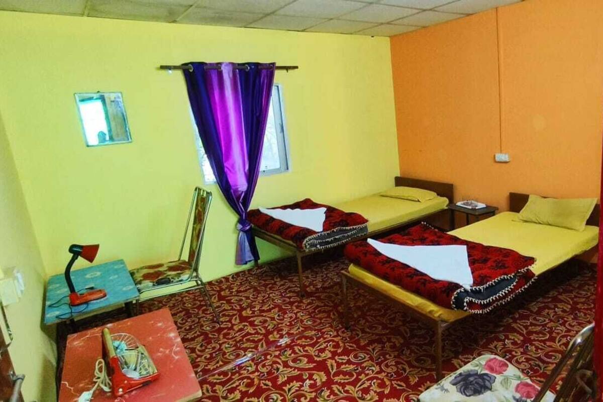 Une chambre avec deux lits, une petite table, une chaise, une fenêtre avec des rideaux violets et un tapis à motifs.