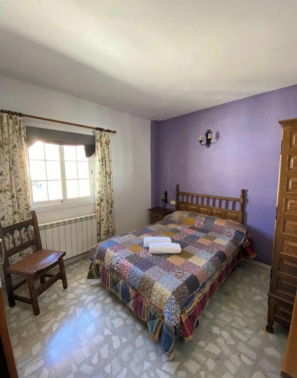 Une chambre à coucher avec un lit en bois, une chaise, une fenêtre avec des rideaux, un radiateur et un luminaire fixé au mur.