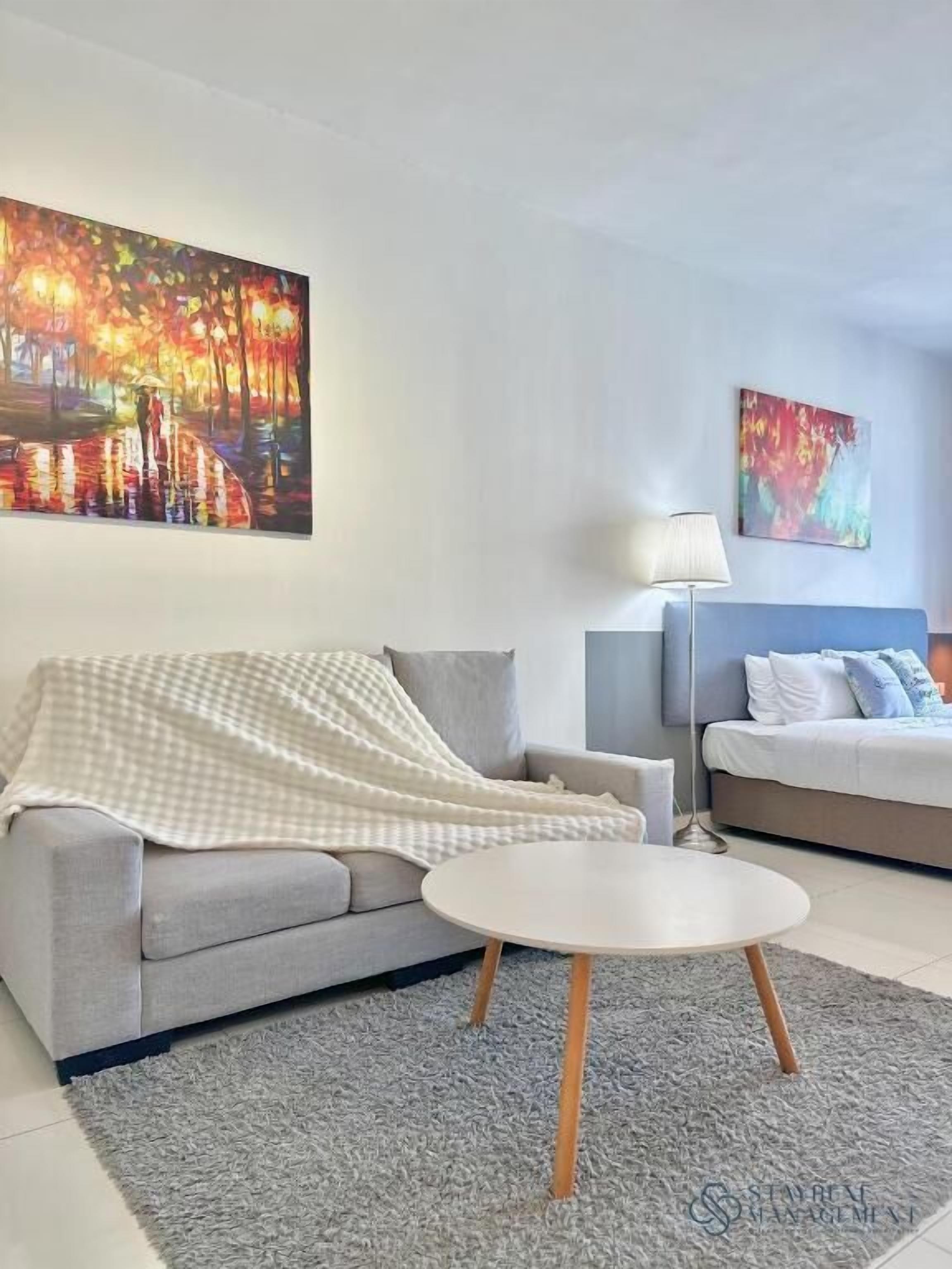 A modern living room with a grey sofa, a round coffee table, and a bed with a blue headboard. There are two paintings on the walls.