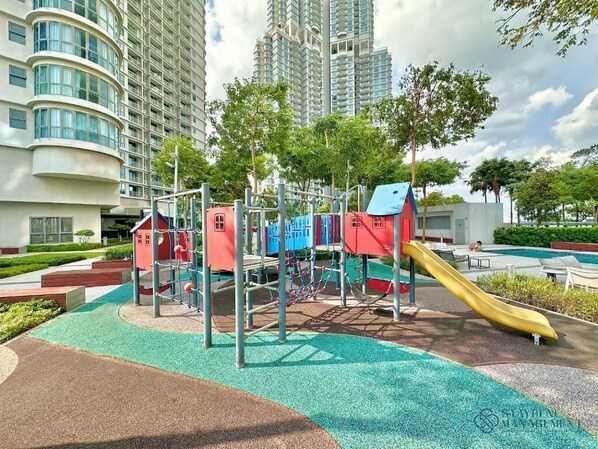 A playground with a slide and climbing structures, surrounded by a landscaped area with trees and a pool in the background. - Teega Suites By Stayrene (Iskandar Puteri)