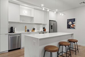 A modern kitchen with white cabinetry, a stainless steel dishwasher, a refrigerator, and a central island with stools. - Minutes from Zilker & Lady Bird | Private Balcony + Pool, Terrace, Dining, Bar (Austin)