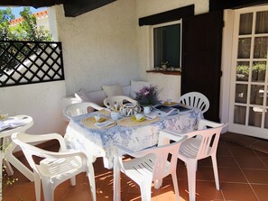A patio with a table set for two, surrounded by white plastic chairs, a white sofa, and a window with a dark frame.
