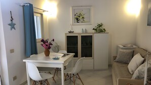 A dining area with a white table, chairs, and a vase of flowers. There is a cabinet with glass doors and a small shelf with decorative items. A couch is visible in the background. - Bouganville Two-Room Apartment 80 m from the Beach with Private Terrace, Garden & AC (Porto Taverna)