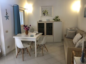 Zona pranzo con tavolo bianco, sedie e un vaso di fiori. Si nota un mobile con ante in vetro e un piccolo ripiano con oggetti decorativi. Sullo sfondo, un divano.