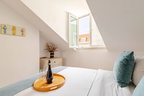 Quarto com clarabóia, cama com almofadas, mesa de cabeceira com um vaso e copos, e janela com vista para edifícios.