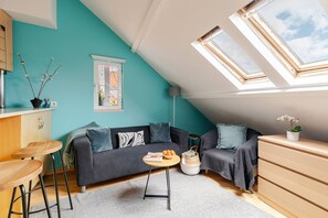 Quarto tipo loft aconchegante com parede em tom de azul-esverdeado, sofá cinza escuro, cômoda de madeira e um claraboia.