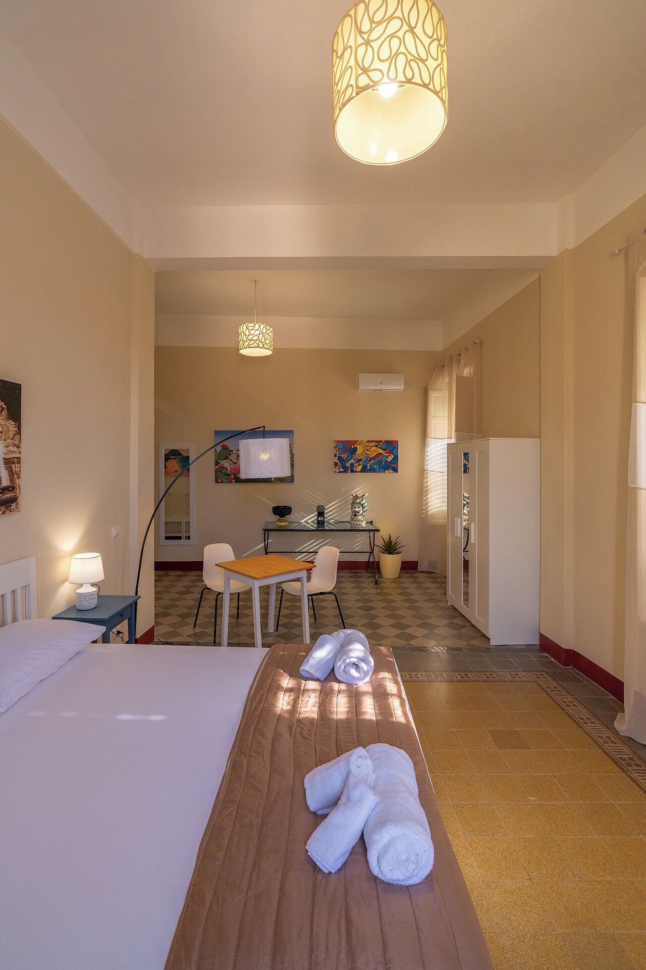 A hotel room with a bed, a wooden table with towels, a desk with chairs, a dining area, and a kitchen.