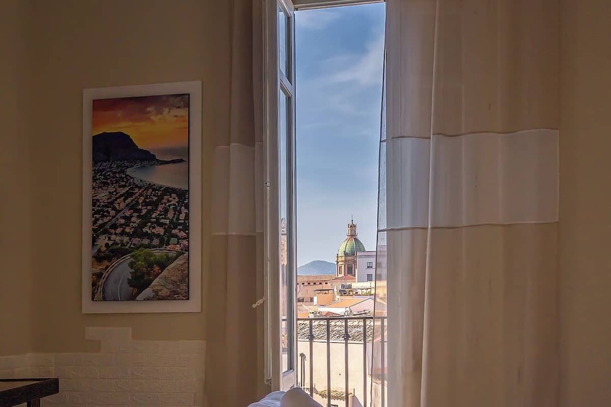 A hotel room with a bed, a painting, and a view of a cityscape through a window.