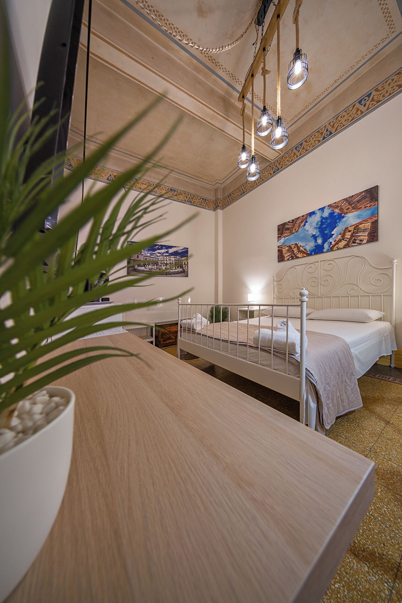 A bedroom with two beds, a wooden desk, a potted plant, and a ceiling with decorative patterns and hanging lights.