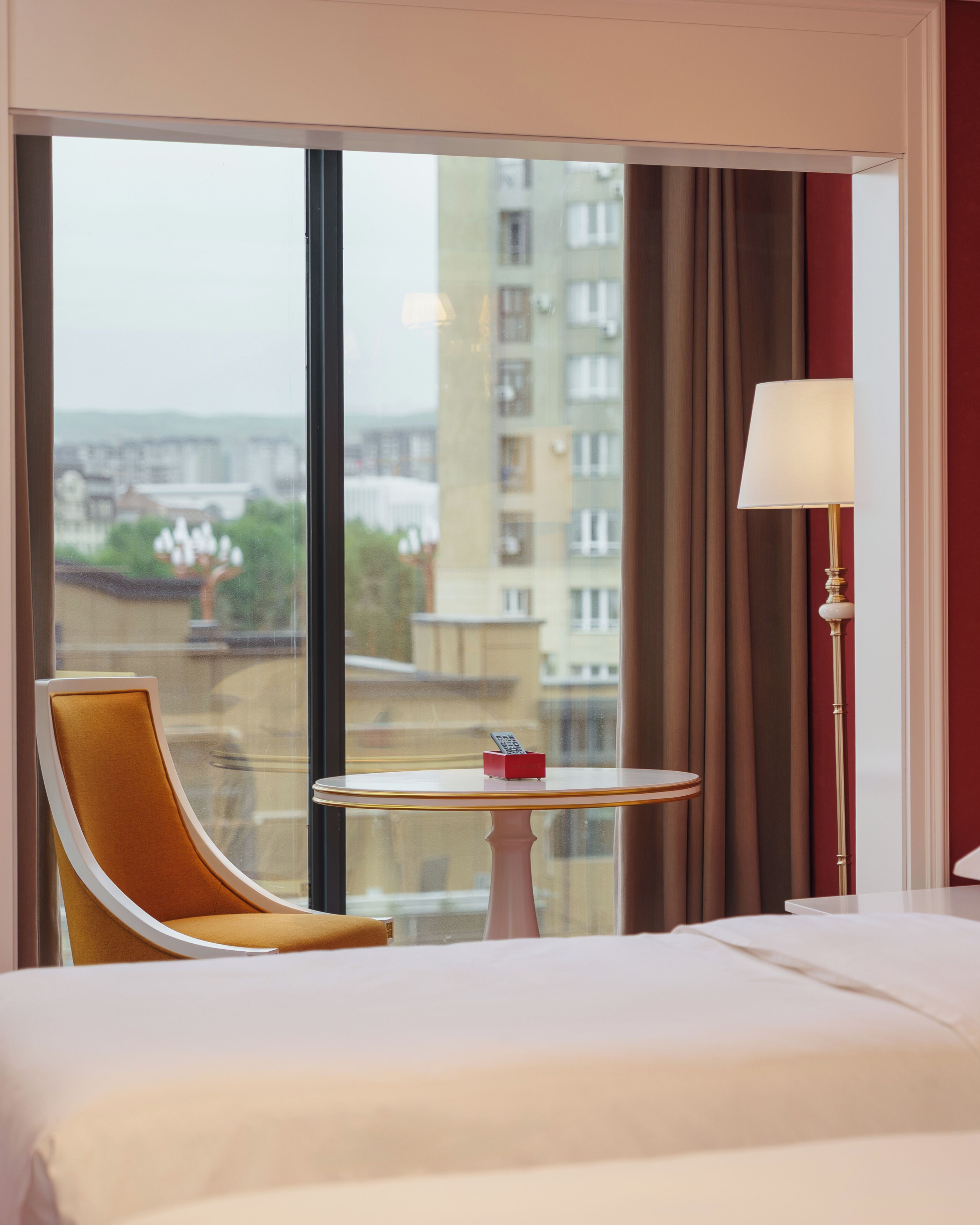 A hotel room with a bed, a round table with a remote control, a chair, and a lamp.