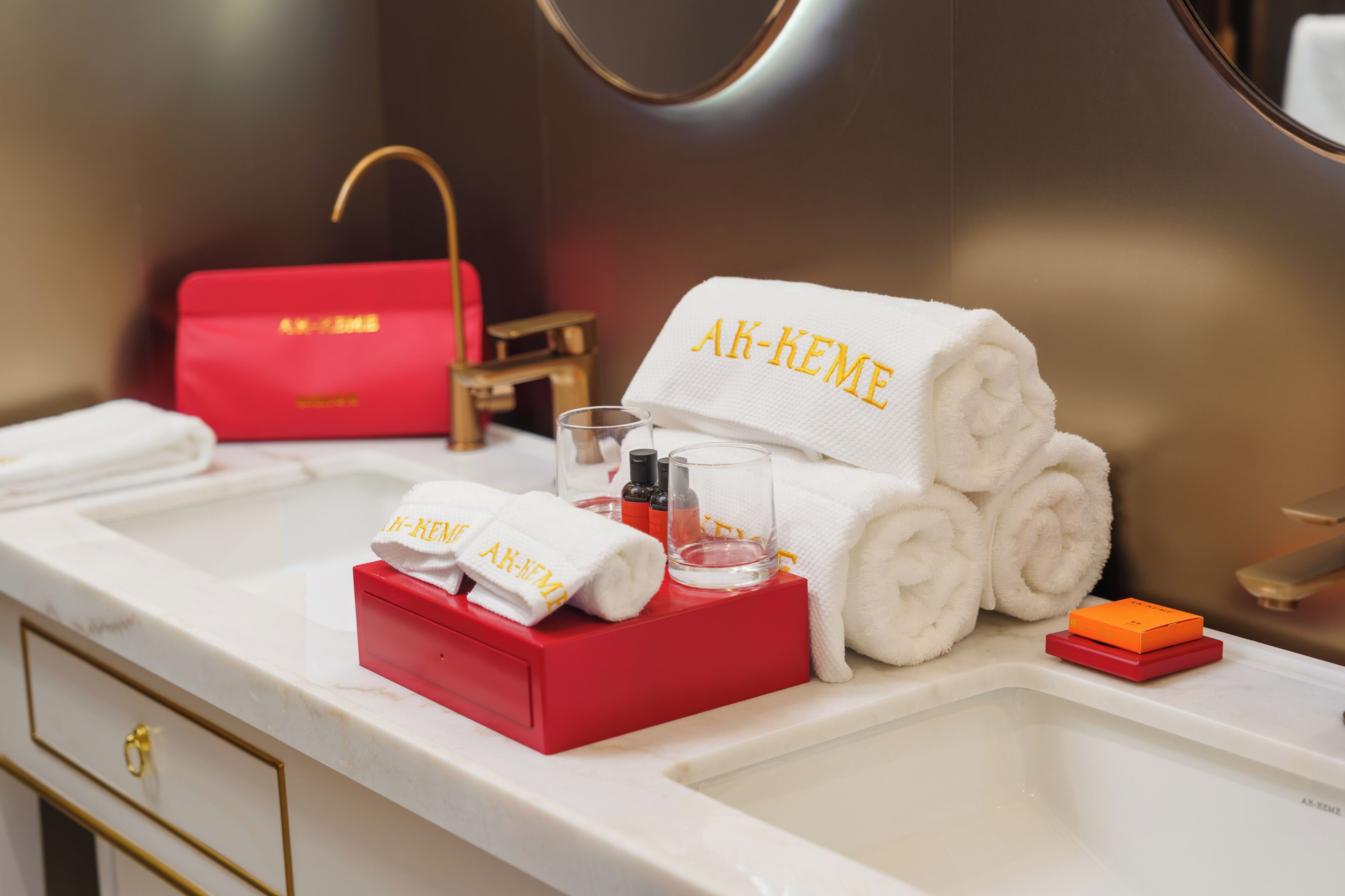Un tocador de baño con espejo, una bolsa de artículos de aseo roja, una toalla blanca con el bordado 'AK-KEME', un vaso de agua y un jabonero rojo.