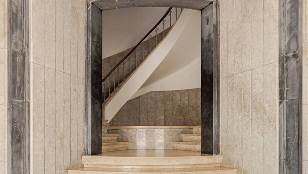 Eine prächtige Treppe mit Marmorstufen führt zu einer Empore, gesäumt von hohen Säulen und einem großen Bogen.