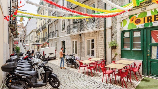 A narrow street with cobblestone pavement, parked scooters, and a café with red chairs and tables. - Trendy Loft Heart Of The City! (Lisboa)
