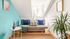 A cozy room with a sofa, a small table with glasses and a bottle, a large plant, and a window with a view of buildings. - Trendy Loft Heart Of The City! (Lisboa)